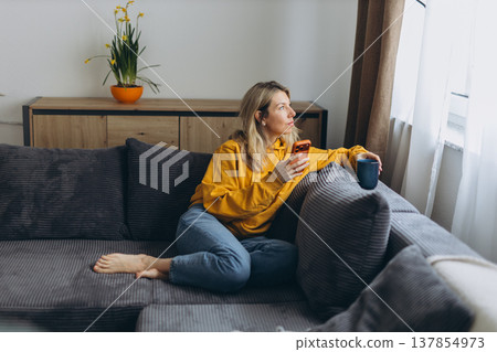Woman holding smartphone and coffee looking outside showing digital distraction and disconnected lifestyle in cozy home setting Woman holding smartphone and coffee looking outside showing digital distraction and disconnected lifestyle in cozy home setting 137854973