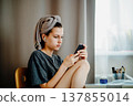 Home scene with a teenage girl looking at her smartphone after a shower. Earphones and a towel on her head add themes of messaging, music and quiet everyday life. 137855014