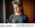 Teen girl standing by the window and looking at her smartphone, with earphones and a towel on her head. Natural home scene about communication, music, morning routine and everyday life. 137855015
