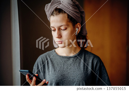 Teen girl standing by the window and looking at her smartphone, with earphones and a towel on her head. Natural home scene about communication, music, morning routine and everyday life. Teen girl standing by the window and looking at her smartphone, with earphones and a towel on her head. Natural home scene about communication, music, morning routine and everyday life. 137855015
