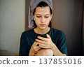 Portrait of a teen girl at home with a smartphone in her hands, earphones and a towel on her head. Natural scene about reading a message, screen focus, music and a quiet personal moment. 137855016