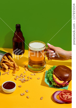 Hand holding beer mug with burger, fries and snacks on bright background with copy space Hand holding beer mug with burger, fries and snacks on bright background with copy space 137855034