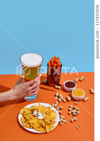 Hand holding beer glass with chips, nuts and dipping sauces on bright background with copy space 137855036