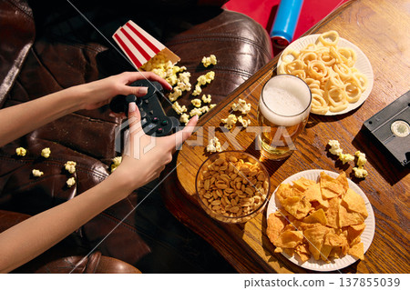 Hands playing video game with beer and snacks on table in cozy setting with copy space 137855039