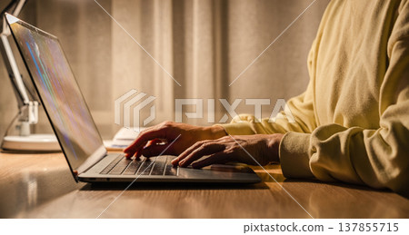 Woman working remotely on a laptop, under warm, soft lighting at a desk. Person typing on laptop keyboard at home at night Woman working remotely on a laptop, under warm, soft lighting at a desk. Person typing on laptop keyboard at home at night 137855715