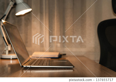 Laptop, lamp, and notepad sitting on a wooden desk, representing remote work, study, and productivity in a home office. Laptop computer illuminating a modern workspace at night 137855717
