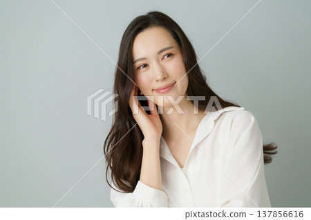 A portrait of a young Japanese woman in a white shirt with her hand on her cheek against a gray background. 137856616