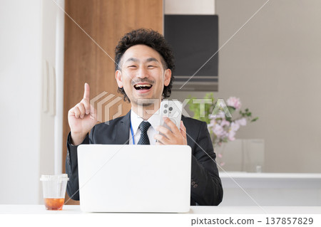 A smiling, bearded businessman in front of his computer, pointing with his smartphone. 137857829