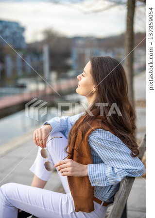 Elegant young woman in old money style sitting on bench near pier in Prague holding sunglasses in early spring. Understated luxury and timeless European femininity concept. Elegant young woman in old money style sitting on bench near pier in Prague holding sunglasses in early spring. Understated luxury and timeless European femininity concept. 137859054