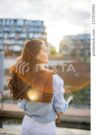 Elegant young woman in old money style standing by riverside in Prague during golden hour, back view portrait. Quiet luxury lifestyle and refined European femininity concept. Elegant young woman in old money style standing by riverside in Prague during golden hour, back view portrait. Quiet luxury lifestyle and refined European femininity concept. 137859060