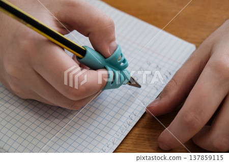 Child practicing handwriting with a special finger grip trainer. Close-up of kid's hand holding pencil correctly. Development of fine motor skills and calligraphy. 137859115
