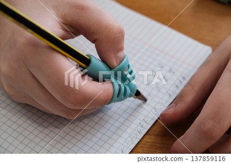 Child practicing handwriting with a special finger grip trainer. Close-up of kid's hand holding pencil correctly. Development of fine motor skills and calligraphy. Child practicing handwriting with a special finger grip trainer. Close-up of kid's hand holding pencil correctly. Development of fine motor skills and calligraphy. 137859116