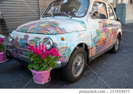 Genoa, liguria, italy. May 18, 2024. Fiat 500 vintage car featuring a painting of colorful houses in cinque terre style, contrasting with bright pink flowers 137859477
