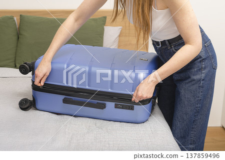 Woman zipping a blue hard shell suitcase on a bed while finishing packing at home. Close-up travel Woman zipping a blue hard shell suitcase on a bed while finishing packing at home. Close-up travel 137859496