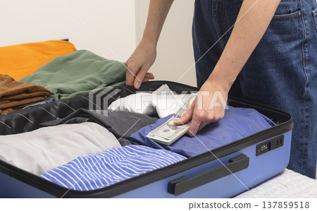 Cash and travel essentials lie beside an open suitcase during packing. Female hands place banknotes Cash and travel essentials lie beside an open suitcase during packing. Female hands place banknotes 137859518