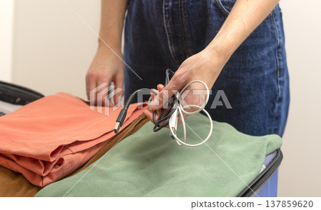 Hands placing charging cables on folded clothes inside an open suitcase during packing at home. Hands placing charging cables on folded clothes inside an open suitcase during packing at home. 137859620