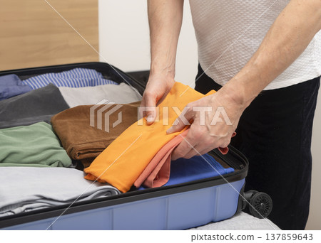 Open luggage on the bed during indoor packing before a trip. Cropped hands fold clothes and place 137859643