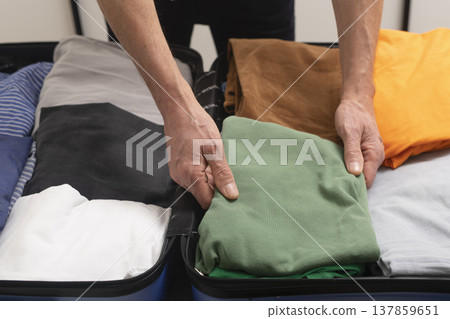 Open suitcase on the bed during indoor packing for a summer escape. Cropped male hands fold clothes Open suitcase on the bed during indoor packing for a summer escape. Cropped male hands fold clothes 137859651