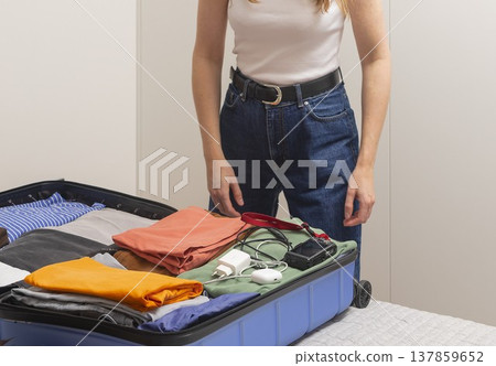 Open suitcase on the bed fills with clothes camera gear and cables during packing. Cropped female 137859652