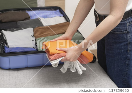 Open suitcase rests on the bed in an apartment during summer packing. Cropped female hands fold Open suitcase rests on the bed in an apartment during summer packing. Cropped female hands fold 137859660