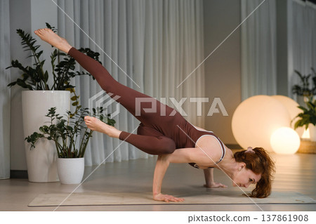 Yoga practice in a studio with a woman performing a challenging pose on a mat during the day with plants and soft lights in the background 137861908