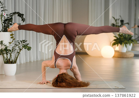 Woman practices handstand yoga pose in a studio with plants and soft lighting during a fitness session in the morning 137861922