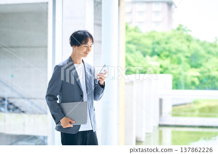 A smiling businessman operating his smartphone while holding a laptop outdoors. 137862226