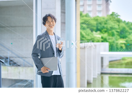 A Japanese man in business casual attire stands with a smile, holding a laptop and a smartphone. 137862227