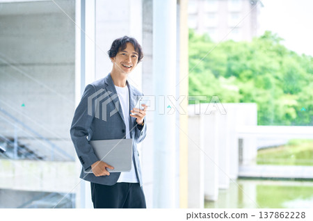 A smiling Japanese businessman in business casual attire, holding a laptop and smartphone. A smiling Japanese businessman in business casual attire, holding a laptop and smartphone. 137862228
