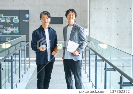Two smiling businessmen in a modern office, each holding a laptop and a smartphone. Two smiling businessmen in a modern office, each holding a laptop and a smartphone. 137862232