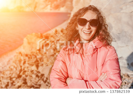 Woman beach vacation smiling in pink puffer jacket and sunglasses on rocky shore during sunset Woman beach vacation smiling in pink puffer jacket and sunglasses on rocky shore during sunset 137862539