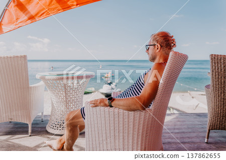 Vacation relax beach man sits in a white wicker chair on a coastal terrace overlooking the sea during a sunny summer holiday Vacation relax beach man sits in a white wicker chair on a coastal terrace overlooking the sea during a sunny summer holiday 137862655