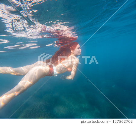 Underwater swimming redhead woman with long hair glides through clear blue ocean water experiencing the serene beauty of nature 137862702