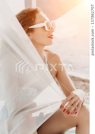 Woman beach vacation sits behind sheer white fabric enjoying a bright sunny day at a coastal resort during summer 137862707
