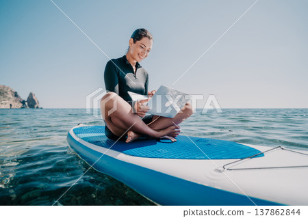 Woman Paddleboard Ocean Reading - A woman sits on a paddleboard in the ocean, reading a book. 137862844