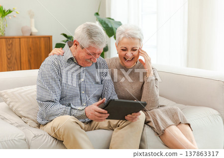 Senior old couple with tablet watching video online. Husband wife spending time together at home. Happy modern mature senior couple using tablet for family entertainment. Old people modern technology Senior old couple with tablet watching video online. Husband wife spending time together at home. Happy modern mature senior couple using tablet for family entertainment. Old people modern technology 137863317