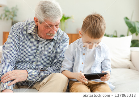 Happy family at home. Old senior man grandfather boy grandson enjoying time together watching video on phone. Good time at home. grandparent and child together. Older generation modern tech usage Happy family at home. Old senior man grandfather boy grandson enjoying time together watching video on phone. Good time at home. grandparent and child together. Older generation modern tech usage 137863318