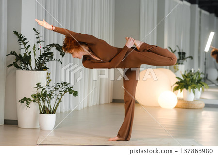 Yoga practice in a studio during daytime with a person balancing in a challenging pose surrounded by plants for wellness and health improvement 137863980