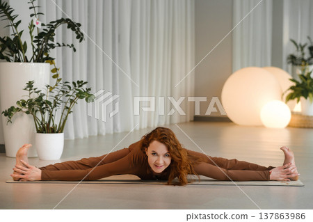 Woman practices yoga in a bright studio surrounded by plants during daytime while stretching her body on a mat with a focused expression Woman practices yoga in a bright studio surrounded by plants during daytime while stretching her body on a mat with a focused expression 137863986