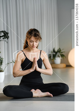Woman practices yoga in a bright indoor space during morning hours while sitting in a cross-legged position with hands in prayer gesture Woman practices yoga in a bright indoor space during morning hours while sitting in a cross-legged position with hands in prayer gesture 137864038
