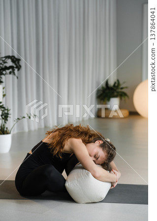 Woman practices yoga in a studio with natural light during a calm session using a cushion for support while stretching and relaxing in a simple pose Woman practices yoga in a studio with natural light during a calm session using a cushion for support while stretching and relaxing in a simple pose 137864041
