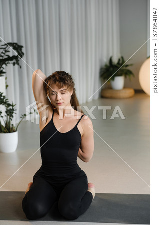 Woman stretches in a serene indoor space with plants while practicing yoga at a wellness studio during daylight hours Woman stretches in a serene indoor space with plants while practicing yoga at a wellness studio during daylight hours 137864042