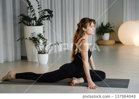 Yoga practice in a studio with plants and soft lighting during the morning hours featuring a woman in a stretching pose on a mat Yoga practice in a studio with plants and soft lighting during the morning hours featuring a woman in a stretching pose on a mat 137864048