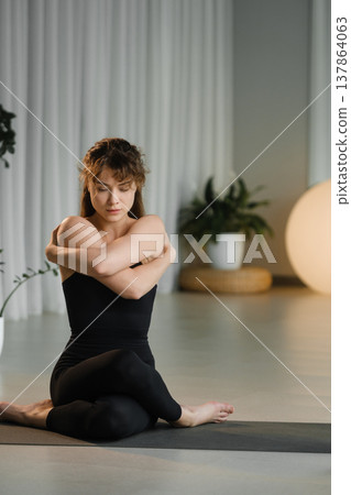 Woman practices yoga indoors while sitting on a mat in a well-lit studio with plants and soft lighting during a morning session 137864063