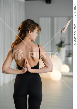 Woman practices yoga in a studio with soft lighting, showing a pose while standing on a yoga mat surrounded by plants in the evening Woman practices yoga in a studio with soft lighting, showing a pose while standing on a yoga mat surrounded by plants in the evening 137864076