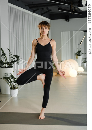 Woman practices yoga in a studio with plants and soft lighting during daytime, showing a balancing pose on one leg with arms raised Woman practices yoga in a studio with plants and soft lighting during daytime, showing a balancing pose on one leg with arms raised 137864079