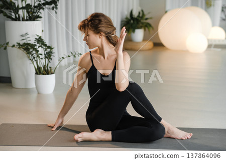 Woman practices yoga in a modern studio with plants and soft lighting during a daytime session focused on flexibility and relaxation 137864096