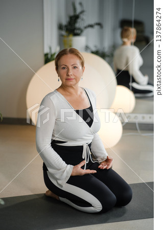 Woman practices yoga in a studio while sitting on a mat surrounded by soft lighting and plants in the background, during the morning hours Woman practices yoga in a studio while sitting on a mat surrounded by soft lighting and plants in the background, during the morning hours 137864274