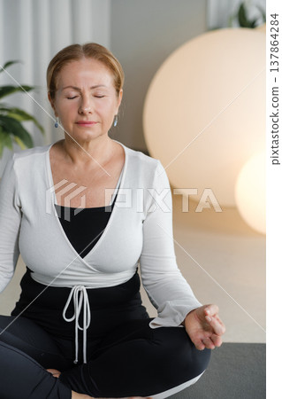 Woman practices yoga in a modern room with a large plant and soft lighting in the evening hours while sitting cross-legged 137864284