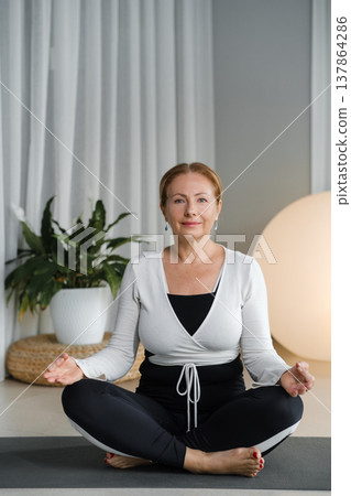 Woman practices yoga in a modern room with a large plant and soft lighting in the evening hours while sitting cross-legged Woman practices yoga in a modern room with a large plant and soft lighting in the evening hours while sitting cross-legged 137864286